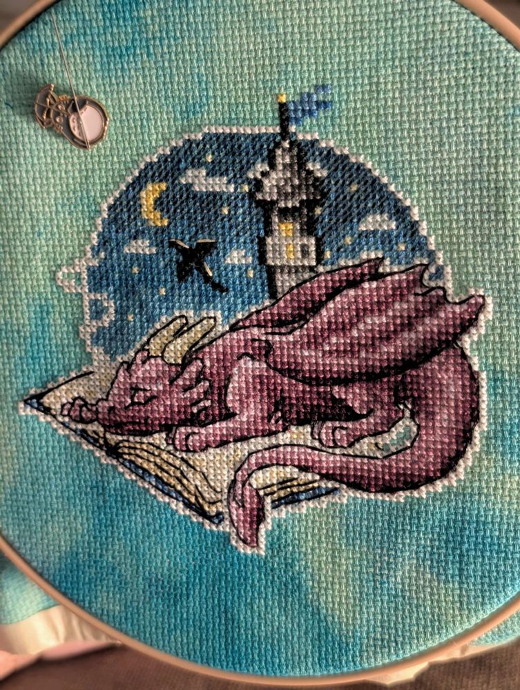 Cross stitch of a cute purple dragon sleeping on an open book, with a flying dragon silhouette and a tower in a night sky background