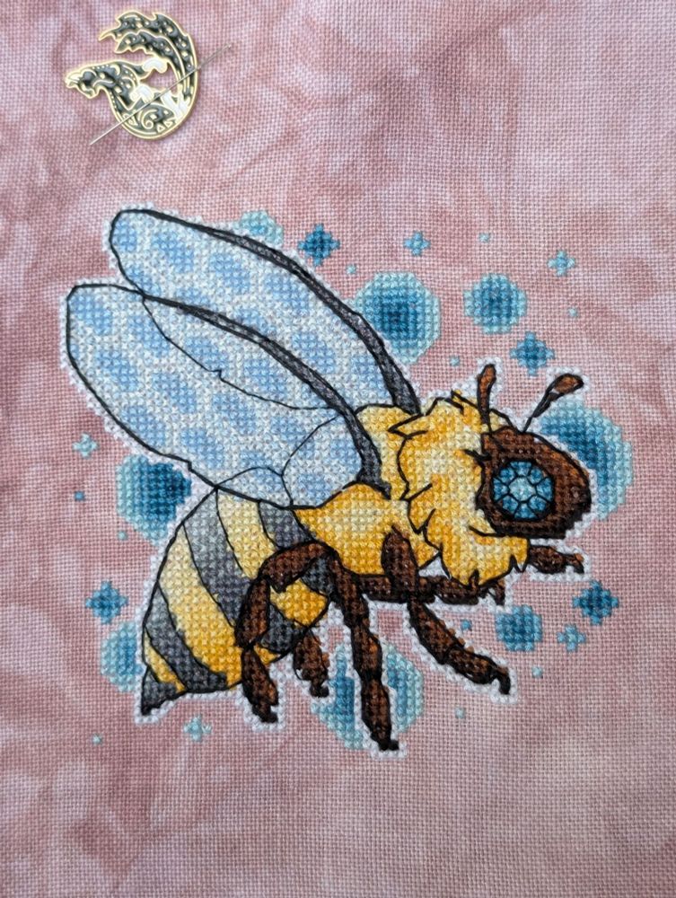 Cross stitched bee with soft blue honeycomb pattern wings and blue gem eyes on mottled mauve linen