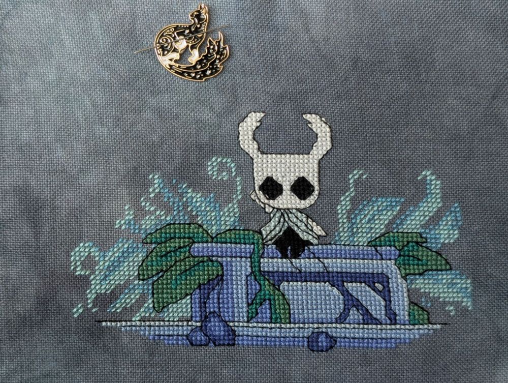 Cross stitch on a mid-tone blue gray linen featuring the Hollow Knight sitting on a blue carved stone with green and teal foliage surrounding