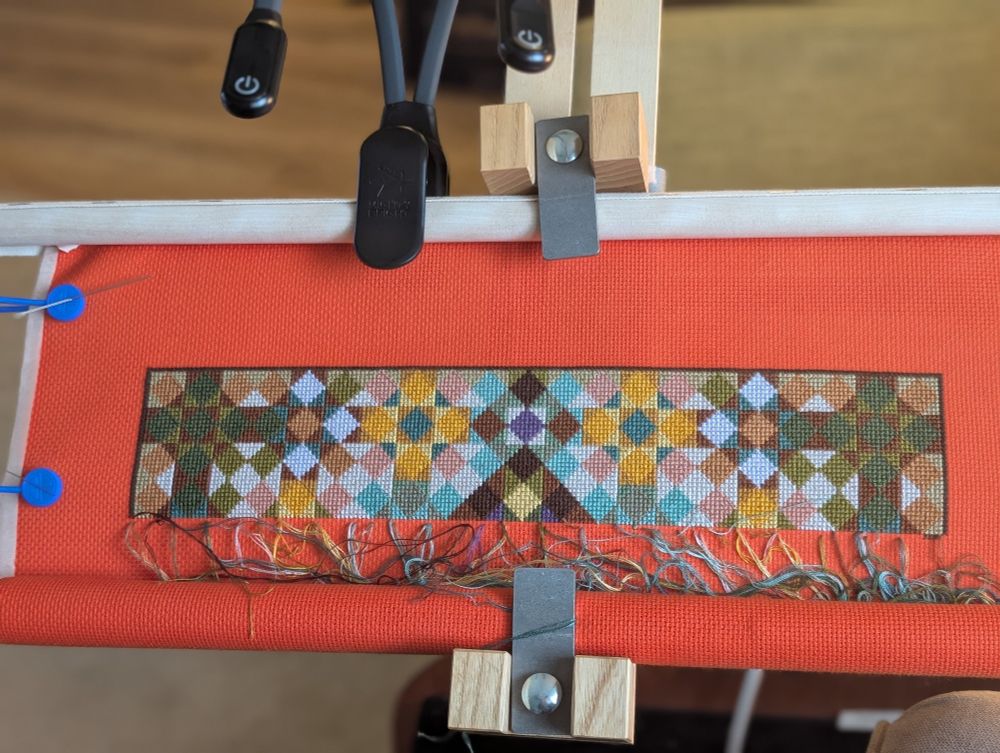 The first few completed rows of a full coverage cross stitch. The pattern is diamonds and triangles making a pattern reminiscent of a kaleidoscope or patchwork quilt. The stitched shapes are in shades of browns, gold, and greens with pops of pale lavender on aida that is traffic cone orange (it looks nicer than that makes it sound, I promise).
