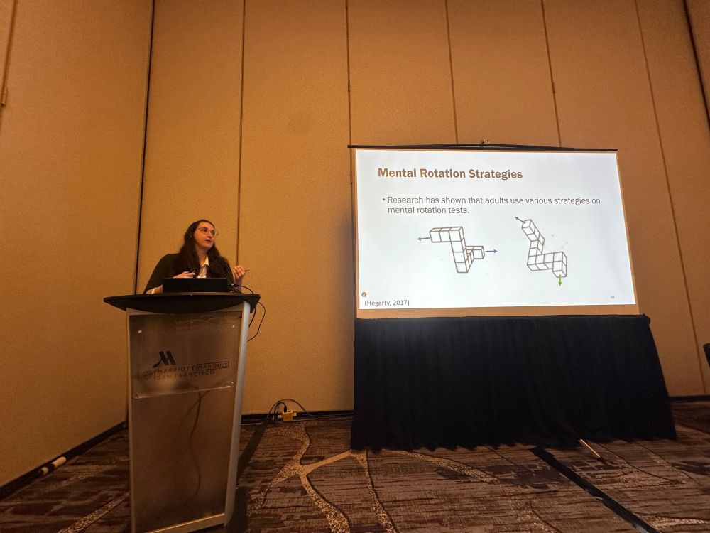 Kiley stands behind podium giving a talk, on the screen it shows “Mental Rotation Strategies” and has two Shepard and Metzler block figures.