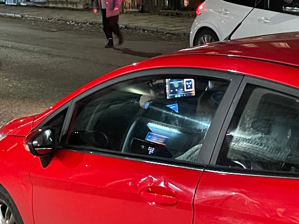 A small red car, looking through the passenger front window. There is a tablet stuck to the windscreen, lit up and being interacted with. It blocks the driver’s view of the road 