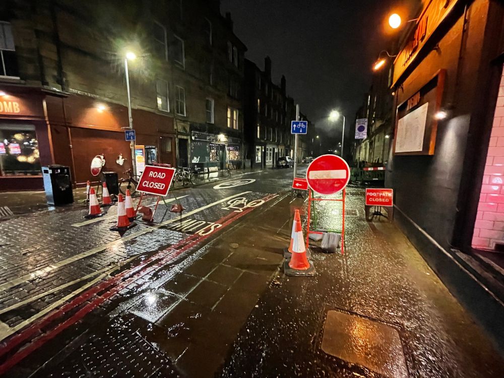 A night scene of a street, with tenements either side. It is wet. There are several temporary no-entry, road closed, pedestrian diversion sign and red traffic cones 