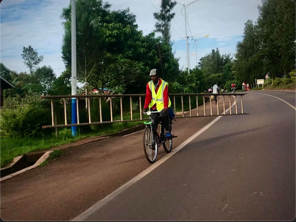 A person rides a bike along the hard shoulder of road, carrying a long ladder at a slight angle. They are wearing a red jacket and hi-viz vest. It looks like an African or Caribbean nation 