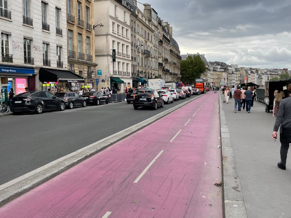 A wide pink coloured bidirectional bike lane. It is empty (but wait a few moments, as the bikes come in bursts). To the right is a wide pavement with several pedestrians. To the left are two lanes for cars and a traffic jam of about 15 cars 