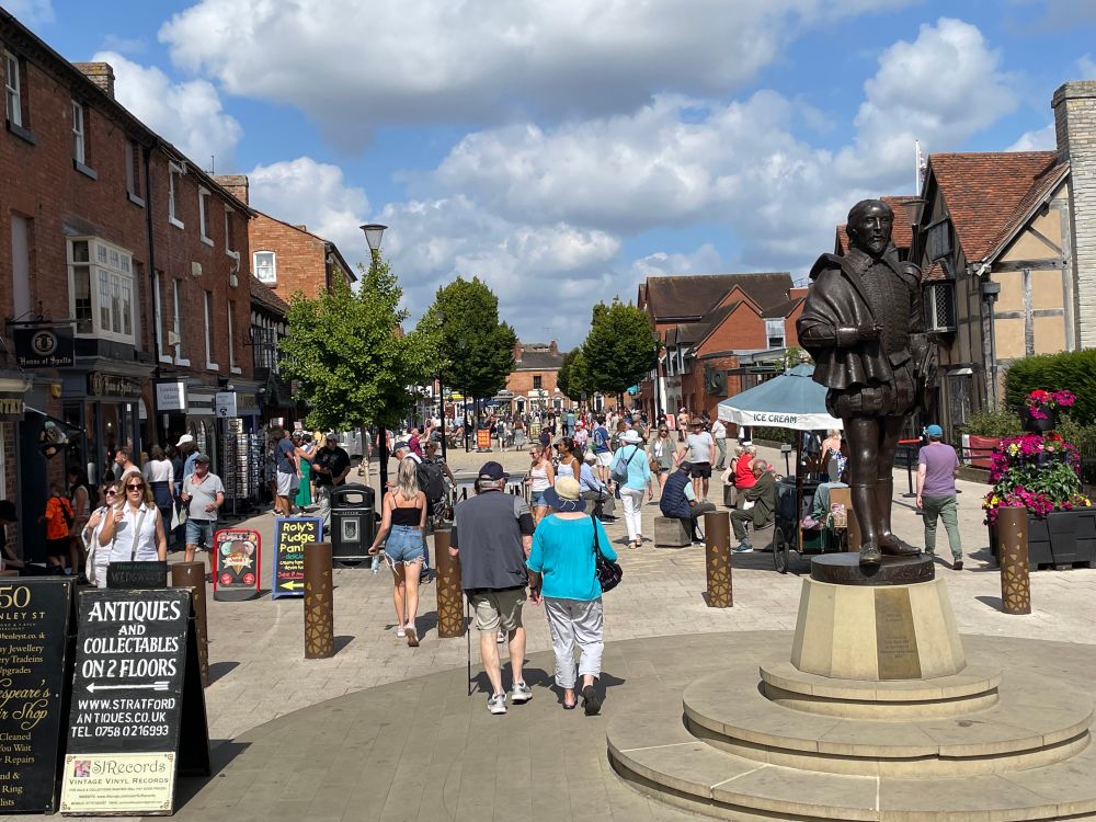 Pedestrianised area thronged with people. A public table tennis table, statue of Shakespeare. Seating buskers