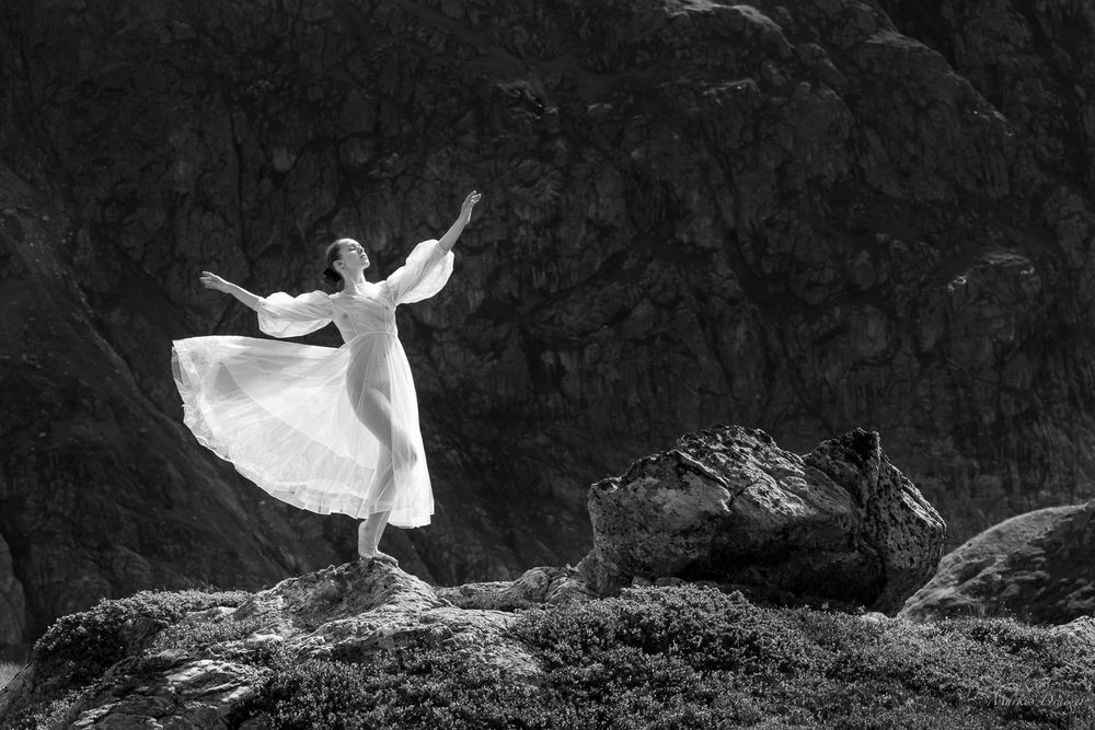 A model wearing a transparent white dress dances in nature on a small rock plate. A dark rocky mountain wall is visible in the back ground. On the right side a bigger rock is lit by a ray of sun light.