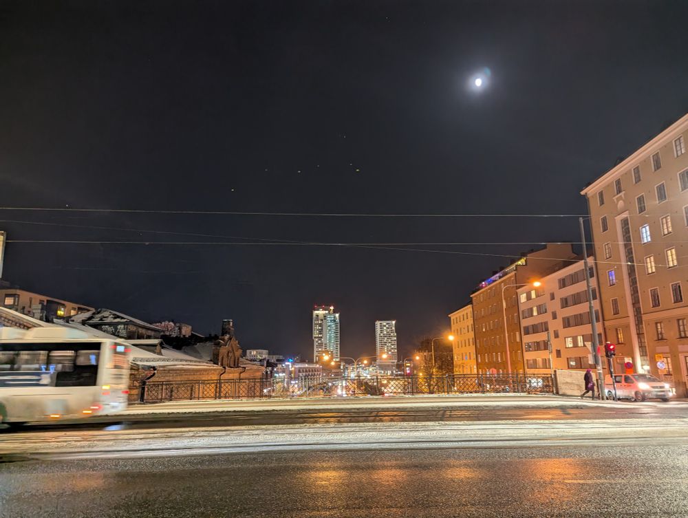 A nighttime cityscape lit with artifical lights and the moon. A view across a city street bridge with light traffic. Beyond the bridge, a bit of highway is visible going into the distance. In the distance, high rise towers against a clear sky