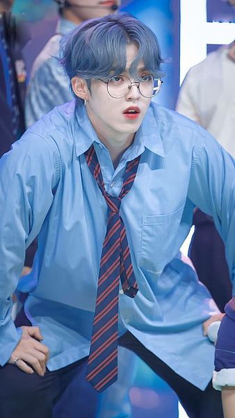 S.coups from Seventeen with his hands on his thighs, slightly in squat position during a stage performance, wearing thin framed glasses, a blue button up shirt, a dark blue and red striped tie worn loosely, his hair dyed a blue grey
