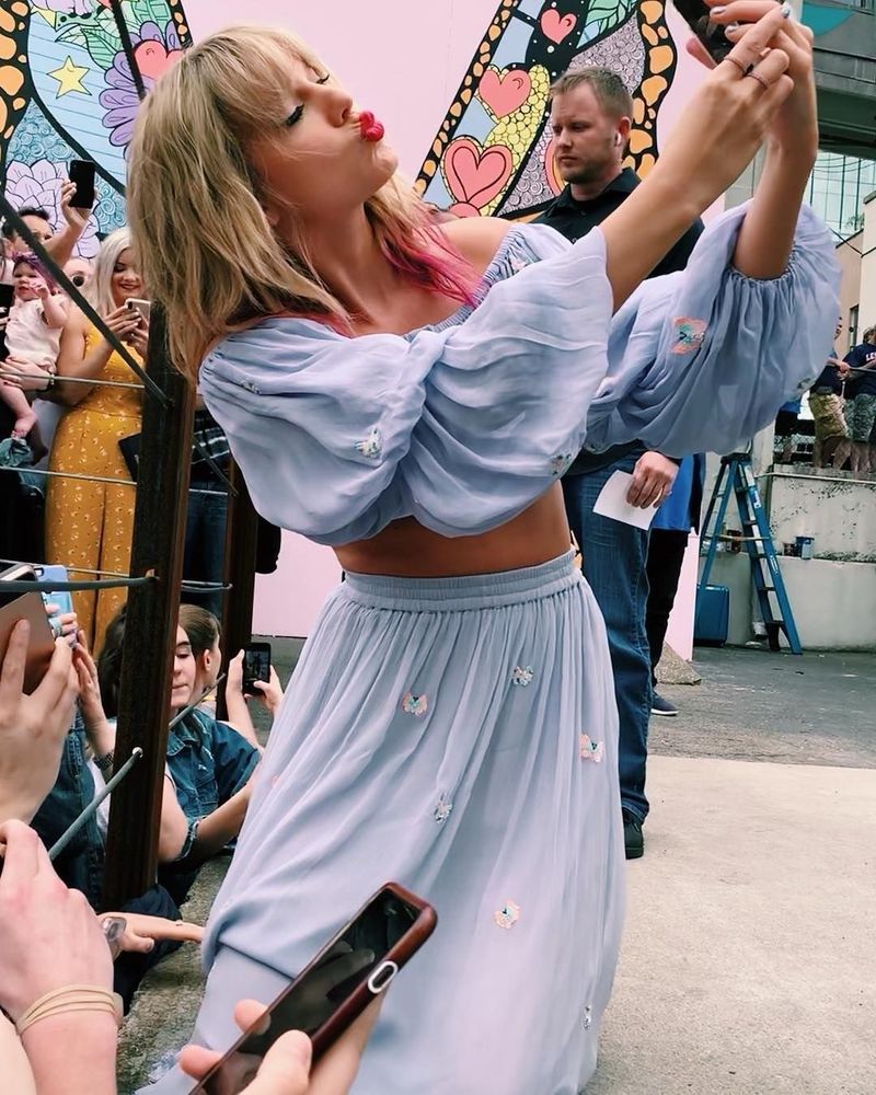 Taylor Swift is in Nashville at the lover mural. She’s wearing a 2 piece outfit with a long sleeve crop top and skirt. It’s a pastel blue colour with butterflies on it. Her blonde hair down with pink tips. She’s making a kissy face as she takes a selfie with the crowd