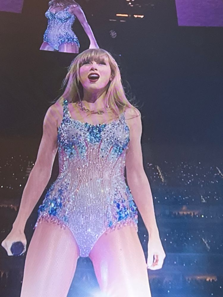 Taylor Swift is at the eras tour in her lover era performing cruel summer. Her blonde hair is done and she is wearing the pink and blue lover bodysuit. She’s holding a mic in her left hand and she’s smiling out to the crowd 
