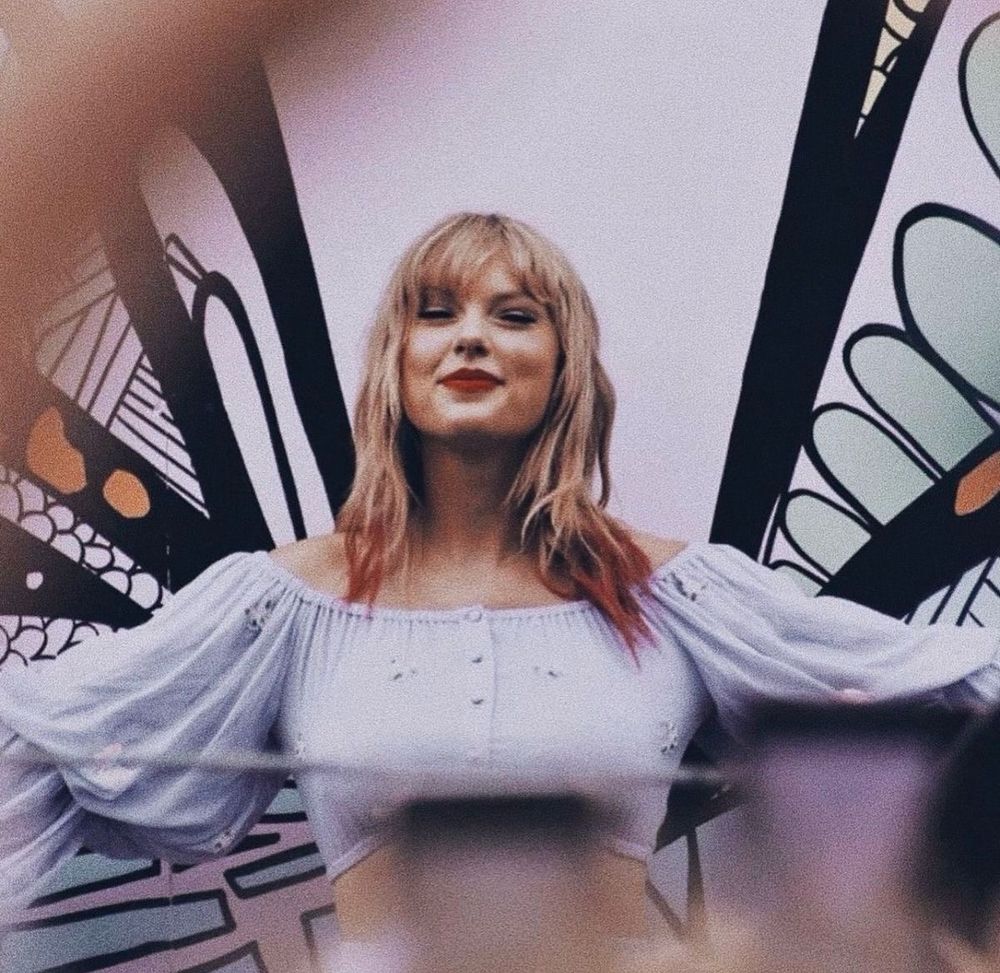 Taylor Swift is in Nashville at the lover mural. She’s wearing a 2 piece outfit with a long sleeve crop top and skirt. It’s a pastel blue colour with butterflies on it. Her blonde hair down with pink tips. She’s looking straight at the camera, her lips red in a closed smile. Her arms spread out to her sides