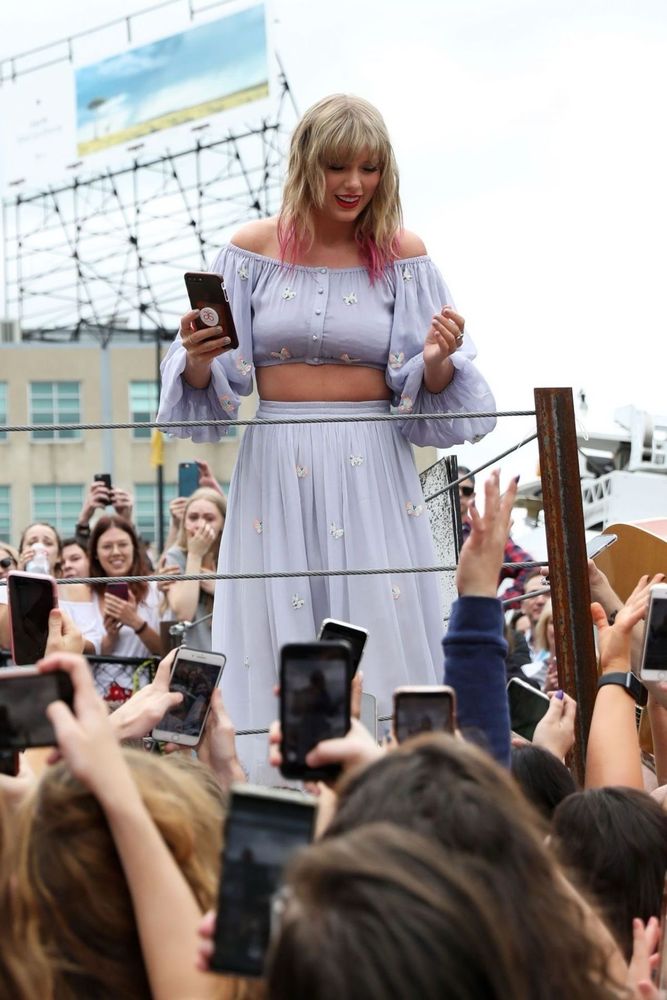 Taylor Swift is in Nashville at the lover mural. She’s wearing a 2 piece outfit with a long sleeve crop top and skirt. It’s a pastel blue colour with butterflies on it. Her blonde hair down with pink tips. There’s a crowd of people below her won’t their phones up in the air taking pics/videos of her