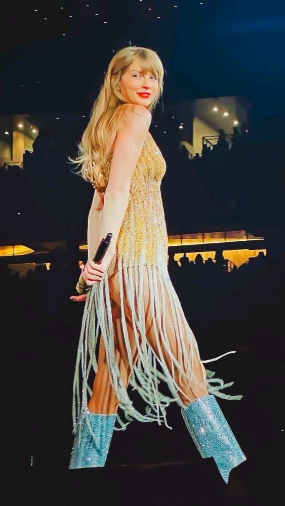 Taylor Swift is performing at the eras tour she’s in her fearless era. She’s wearing the gold spaghetti dress and silver knee high boots. Her long blonde hair flowing behind her back as she looks to the side into the crowd her red lips are smiling and she’s carrying the mic in her right hand behind her back