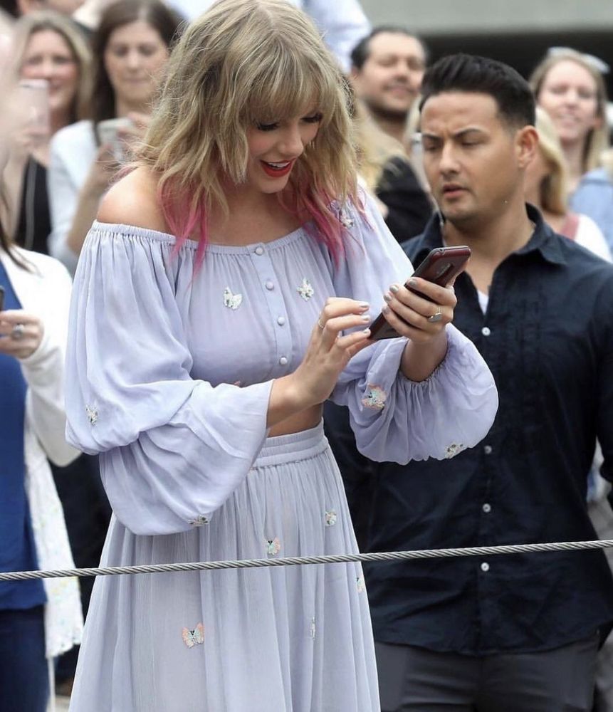 Taylor Swift is in Nashville at the lover mural. She’s wearing a 2 piece outfit with a long sleeve crop top and skirt. It’s a pastel blue colour with butterflies on it. Her blonde hair down with pink tips. She’s smiling and looking down at a phone 