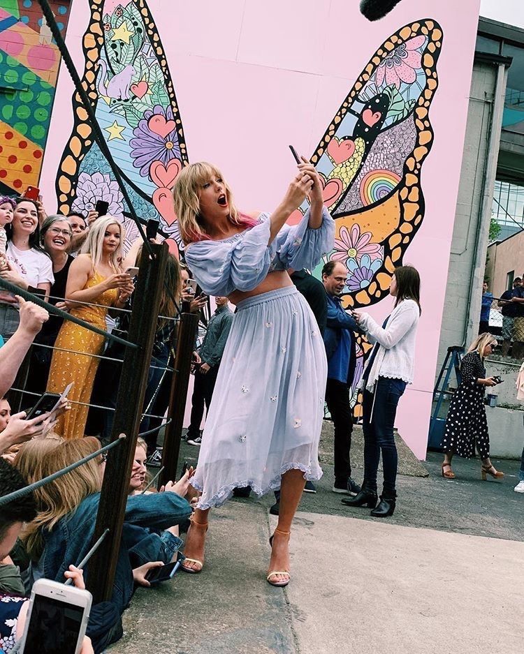Taylor Swift is in Nashville at the lover mural. She’s wearing a 2 piece outfit with a long sleeve crop top and skirt. It’s a pastel blue colour with butterflies on it. Her blonde hair down with pink tips. Behind her is the butterfly mural. She’s leaning to her right holding up a phone taking a selfie with the crowd
