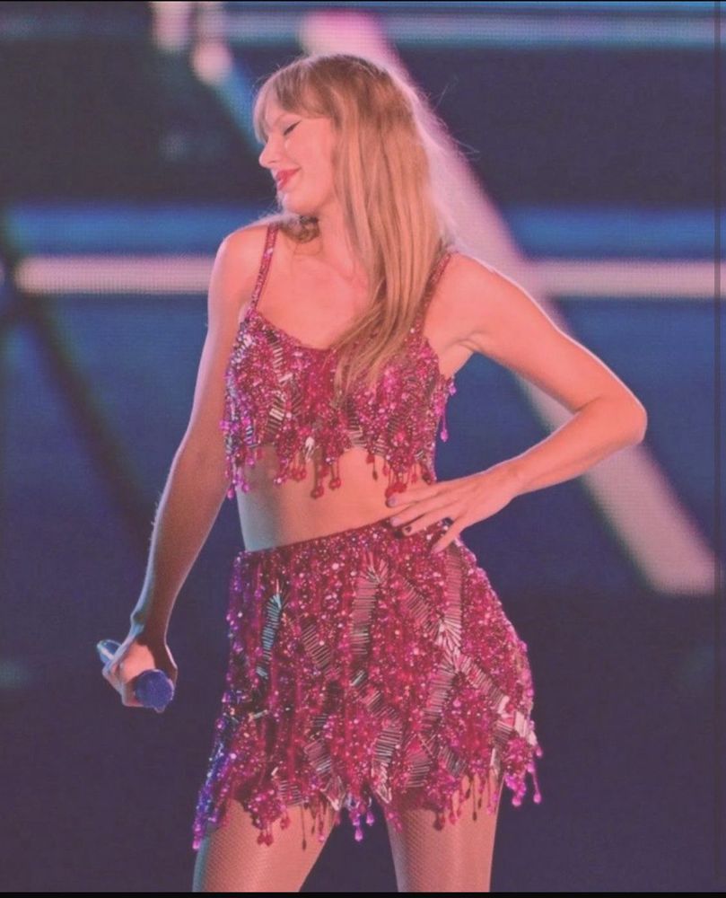 Taylor Swift is performing at the eras tour, in her 1989 era. She’s got her left hand on her hip and her right hand at her side holding the mic. She’s wearing the original pink 2 piece outfit. Her long blonde hair down, her eyes closed and a closed lip smile on her face 
