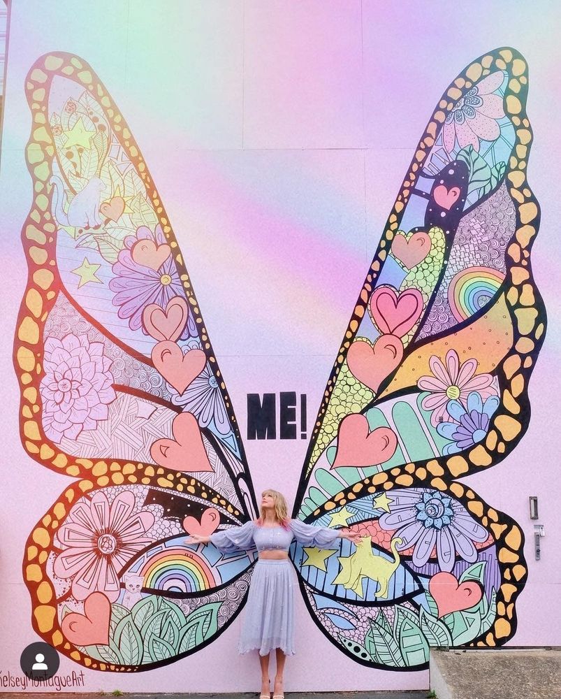 Taylor Swift is in Nashville at the lover mural. She’s wearing a 2 piece outfit with a long sleeve crop top and skirt. It’s a pastel blue colour with butterflies on it. Her blonde hair down with pink tips. Her arms spread out either side of her. Behind her is the beautiful butterfly mural full of colour. On the middle of the wings it reads “ME!” 