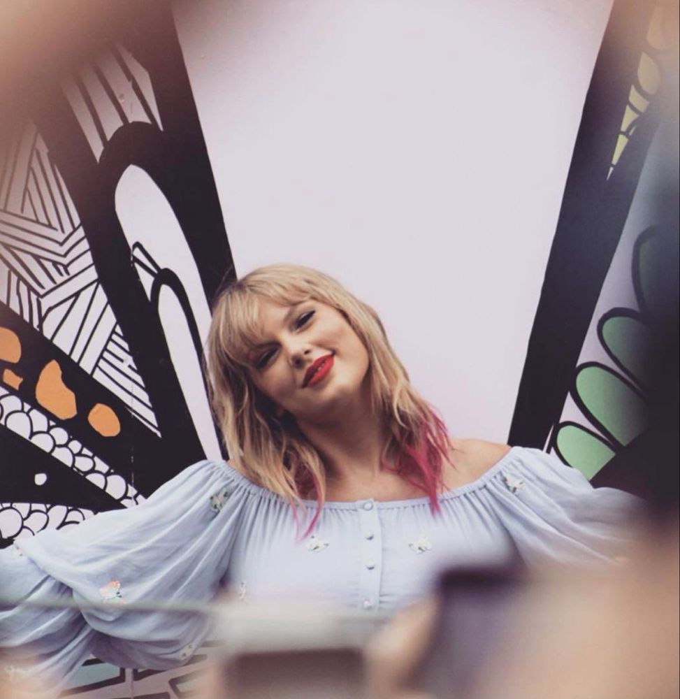 Taylor Swift is in Nashville at the lover mural. She’s wearing a 2 piece outfit with a long sleeve crop top and skirt. It’s a pastel blue colour with butterflies on it. Her blonde hair down with pink tips. Her head tilted to the side and she smiles at the camera. Her arms out at her sides