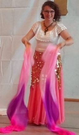 Une femme en tenue de danse : jupe longue rose, large ceinture de sequins or, un haut blanc manches courtes en dentelle . Elle tient des éventail voile fushia, violet, rose. 