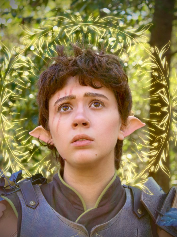 Sophie is dressed in cosplay as Orym, a halfling from critical role’s third campaign. She is seen from the shoulders up in green and brown leather armor, with short brown hair and pointed ears. She’s looking upward, with a serious expression and a scar across her right eye. There is a drawn faintly glowing halo of leaves and flowers surrounding her head and the photo has a dreamy, warm-toned cast to it.  