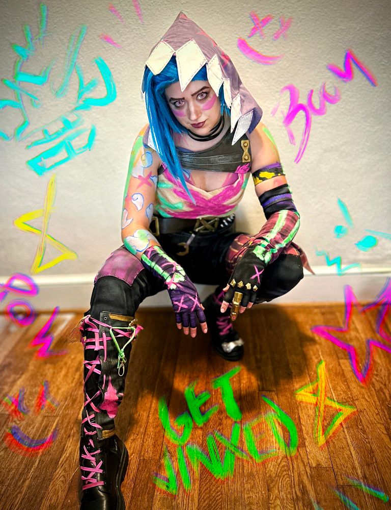 Sophie is dressed at Jinx from Arcane, wearing her outfit from Act 3 of Season 2. She is crouched with one leg forward, looking into the camera. There are drawn symbols surrounding her. 