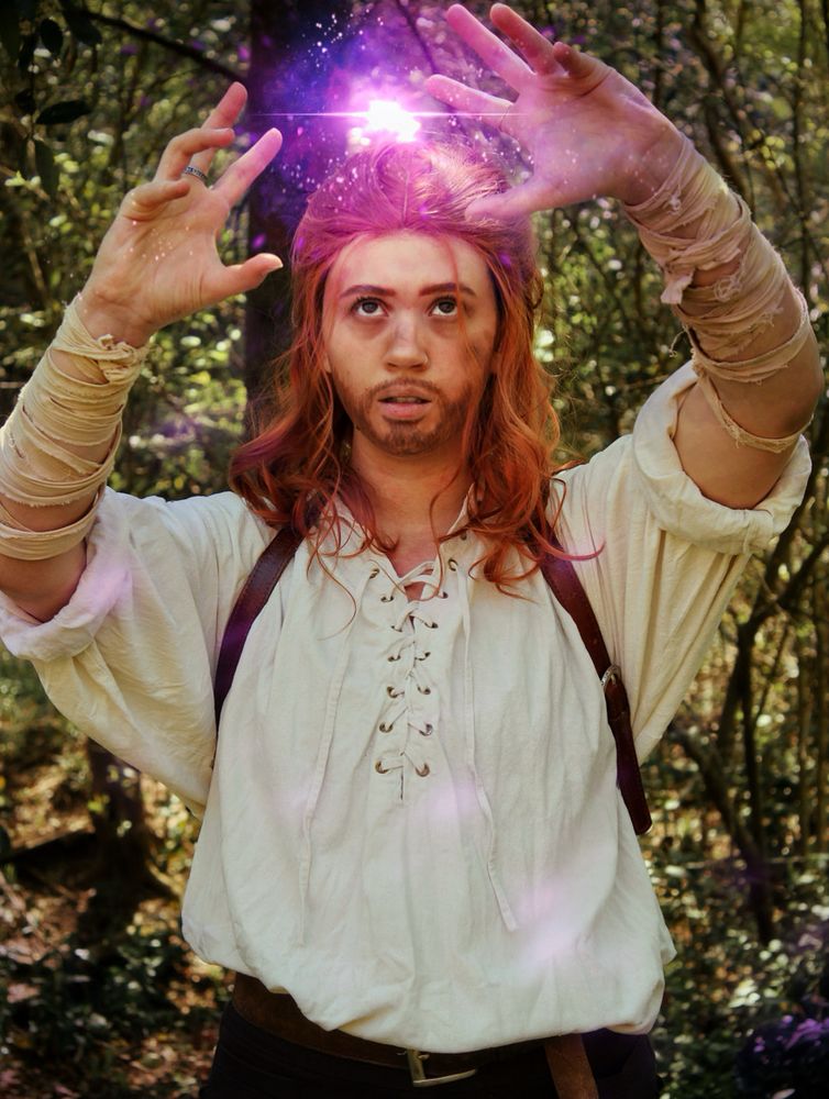 Soph is dressed in cosplay as Caleb Widogast from critical role’s second campaign. They are in a loose shirt with a leather harness across the shoulders, with messy red hair and a beard, and dirt on their face. Their hands are up, as if casting a spell, with purple energy gathering in the center. They are standing outside in a forest. 