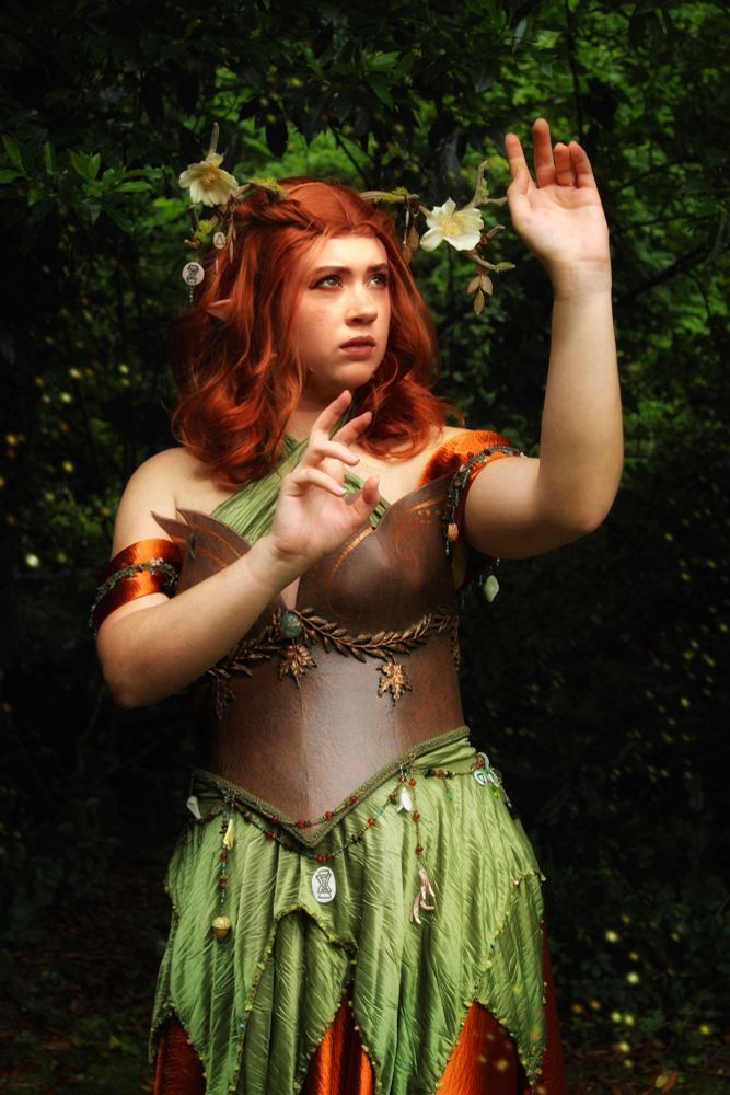 Sophie is dressed in cosplay as Keyleth, from Critical Role’s first campaign. She is in a leather bodice with an orange and green dress, and decorated antlers. She is holding her hands up as though casting a spell, and looking into the distance. 