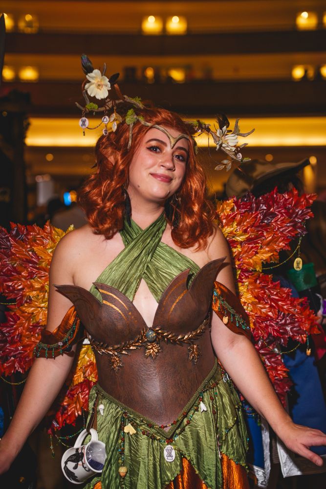 Sophie is dressed in cosplay as a fae version of Keyleth from Critical Role and the Legend of Vox Machina, with a green and orange dress and brown corset. She has short red hair and antlers, and wings made from maple leaves. 
