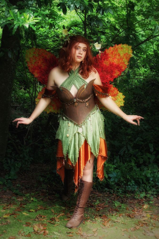 Sophie is dressed as a fae version of Keyleth from Critical Role. She has autumn leaf wings and is standing in a forest. 