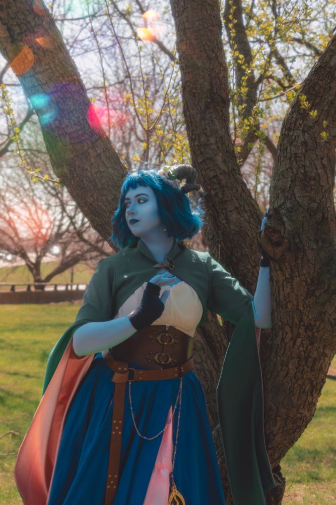 Sophie is dressed in cosplay as Jester Lavorre from critical role’s campaign 2. She is in blue body paint dressed as the outgoing tiefling cleric, and is standing outside looking into the distance with her hand on a tree. The sunlight is behind her and a breeze is blowing her flowing sleeves in the wind. 