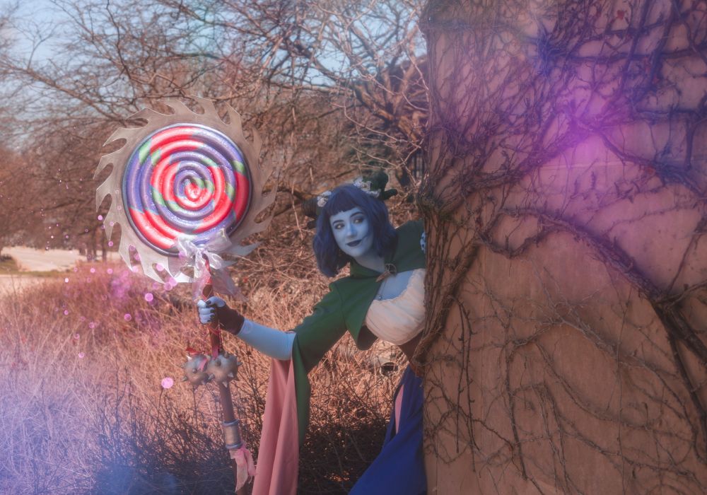 Sophie is dressed in cosplay as Jester Lavorre, a blue tiefling from Critical Role’s second campaign. She is holding a large faux lollipop and peeking around a vine-covered wall