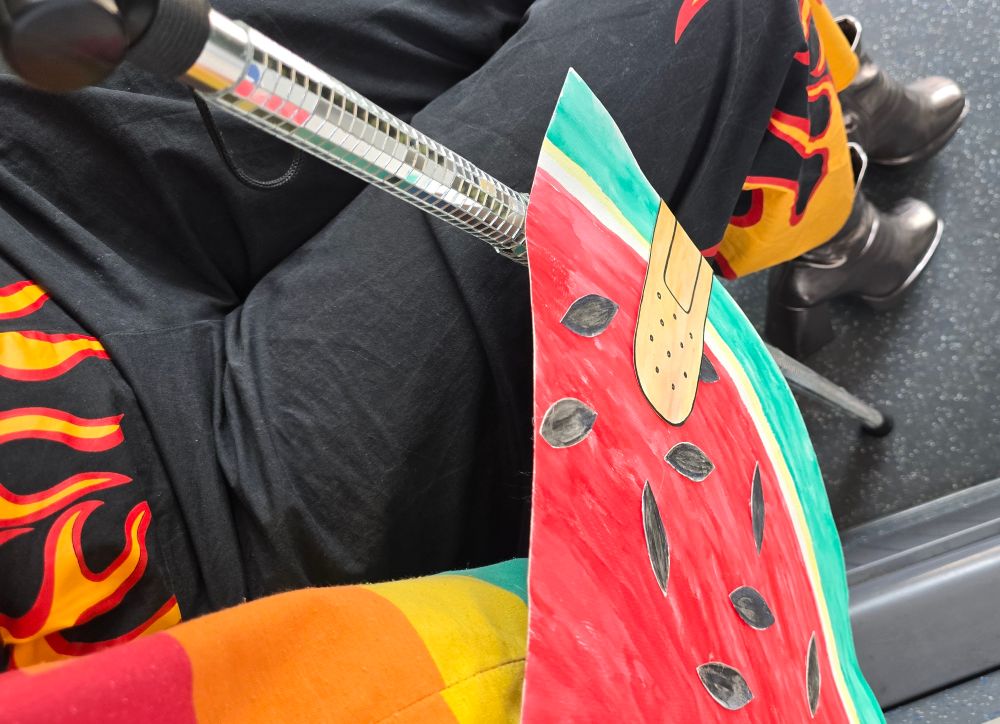 Pantalon noir motif flammes, canne boule à facettes et totebag arc-en-ciel auquel est accroché une pastèque géante en carton