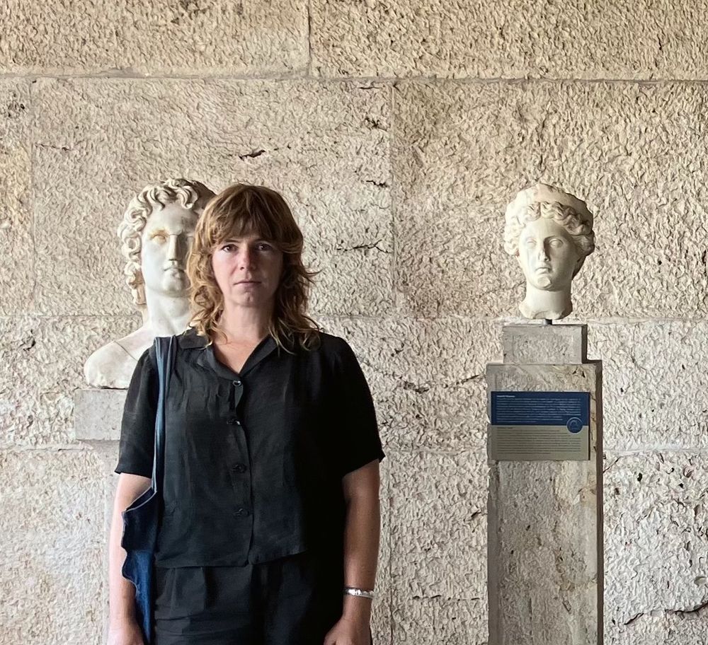 Photo of kelly standing in front of some Greek statuary. One of the figures is behind her to the left, like an echo.