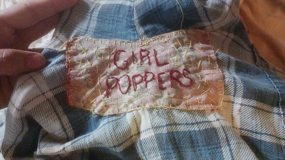 In the pit of a generic faded blue and white flannel, a wrongside-out patch of fabric attached with yellow thread in a blanket stitch and little stars, there's a big maroon embroidery of the words, "GIRL POPPERS"