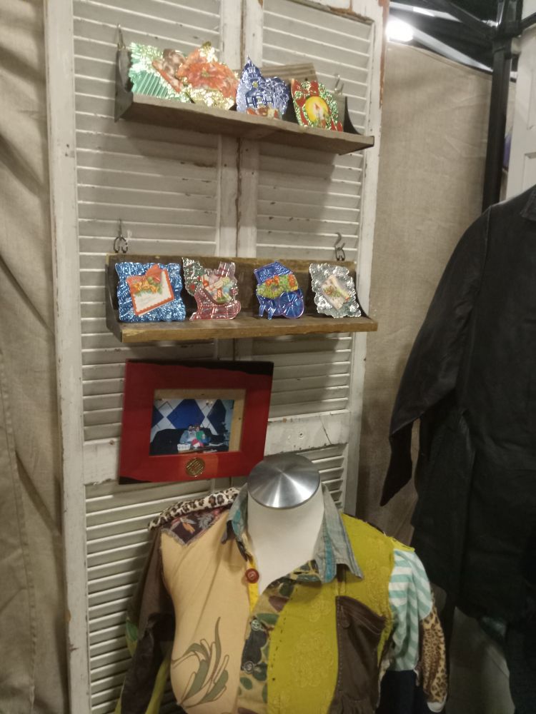Beneath a very pomo frame with an enron logo is a mannequin in a shirt made of mismatched fabric scraps. Above is a pair of rough wood shelves with 1940s foil Christmas cards arranged on them. You cab see a leather jacket to the side