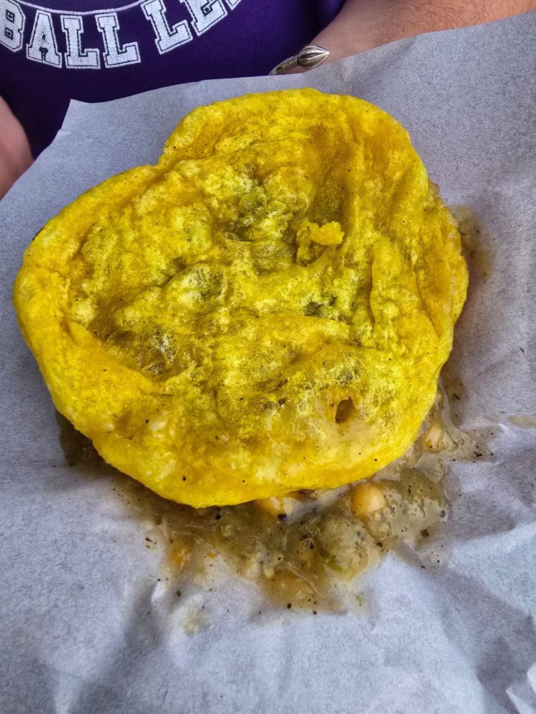 A freshly assembled Trinidadian “doubles” sits on a sheet of wax paper—fluffy, golden‑yellow bara (fried flatbread) draped over curried chickpeas and pepper sauce that peek out around the edges. Flecks of green seasoning and turmeric tint the bread, while a few chickpeas and thin gravy have pooled beneath. In the background, part of a person’s torso in a purple T‑shirt and a silver bangle on their wrist frames the street‑food snack.
