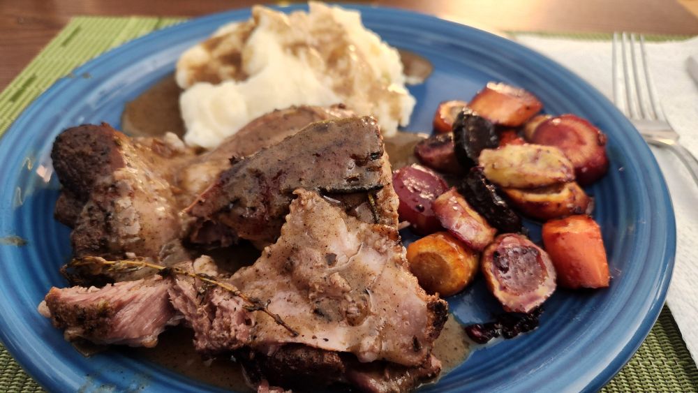A blue dinner plate holds sliced roasted lamb shank with rich gravy, creamy mashed potatoes, and a colorful medley of roasted carrots in shades of orange, yellow, and purple. The table is set casually with a napkin, fork, and placemats