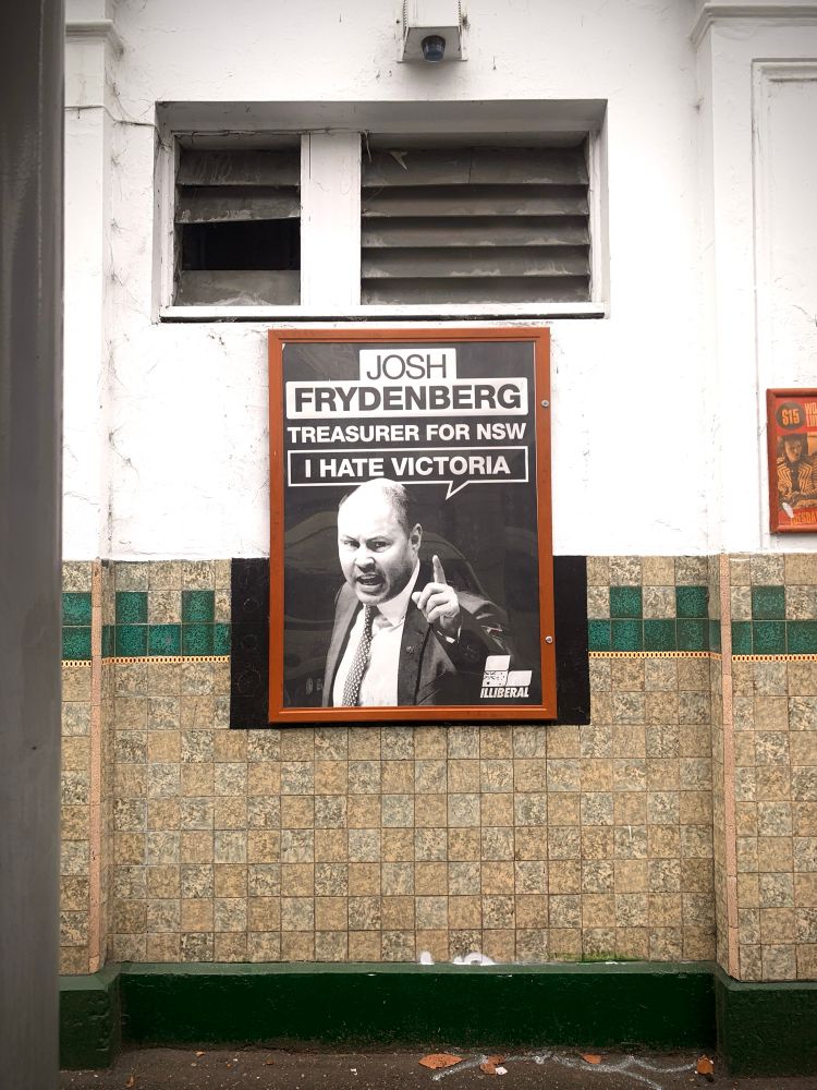 Poster in a wooden frame on the tiled wall Victorian era wall of a pub on Gertrude St Fitzroy. Bottom half features a mid shot photo of Josh Frydenberg taken from about 45 degrees above. It looks like he is speaking in parliament, he is pointing his finger. The title text - top line: JOSH, 2nd line: FRYDENBERG, 3rd line: TREASURER FOR NSW. There is a text bubble under the top text. It points from above his pointing finger near his head that says “I HATE VICTORIA” 