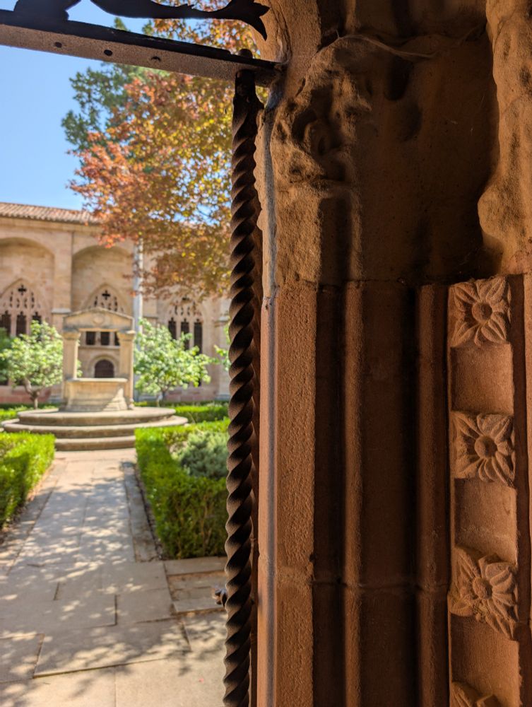 Ventana de piedra trabajada abierta al claustro de la catedral de Sigüenza. Pozo en el centro, jardín de setos y árboles, arcos de piedra y el cielo azul.