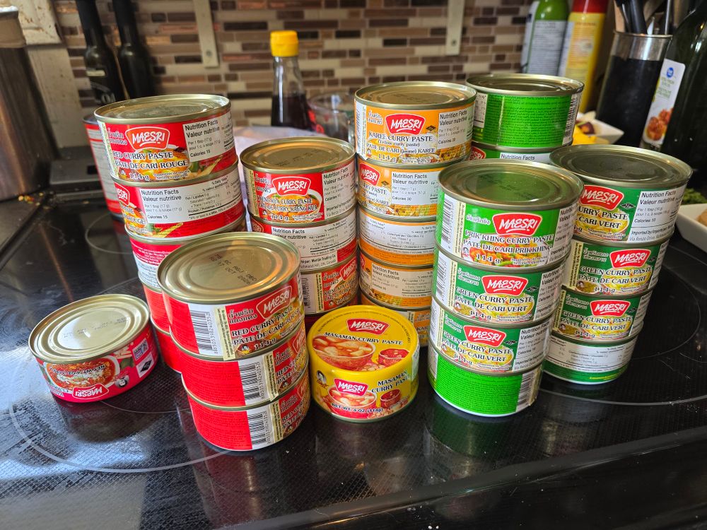 A couple dozen brightly coloured little cans of different flavours of maesri Thai curry paste stacked on a stove top.