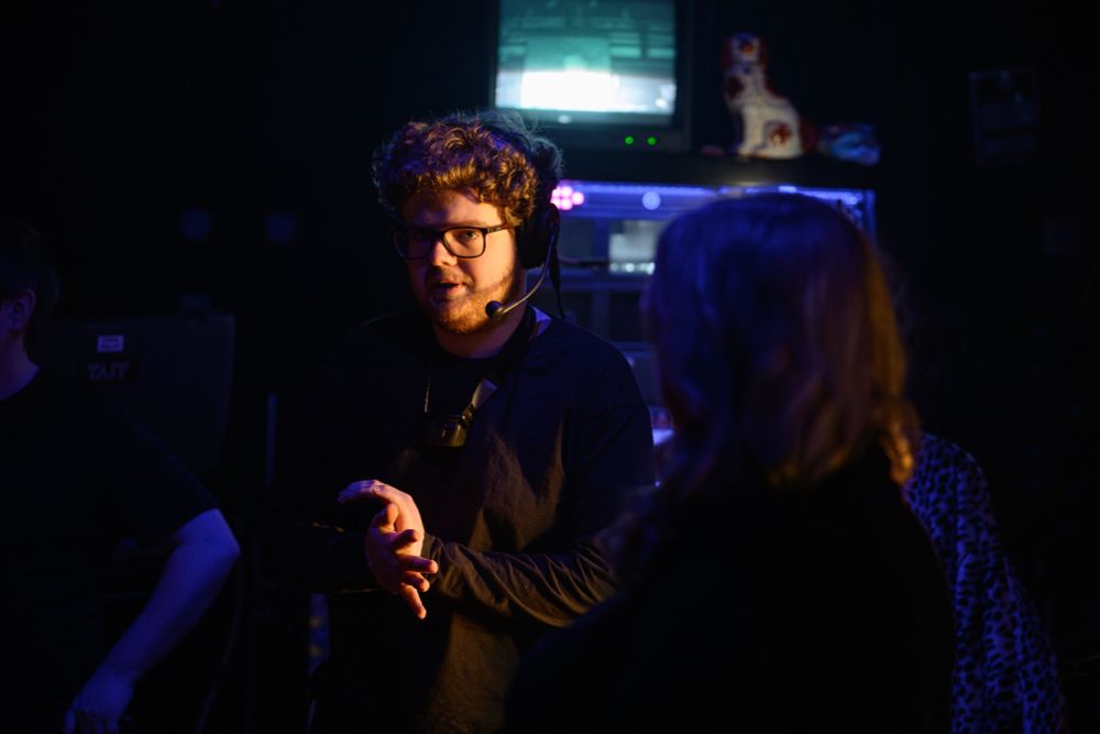 A stage crew member wearing a headset and glasses is talking to a colleague in a dimly lit backstage area with equipment and monitors in the background.