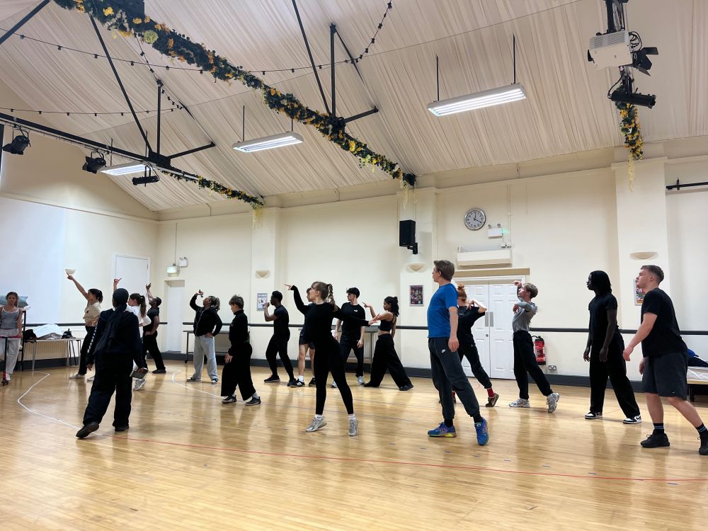 A group of performers in a spacious studio, practicing synchronized dance moves. Some are standing with arms raised, while others are mid-motion. The studio has high ceilings and floral garlands hanging from the beams.