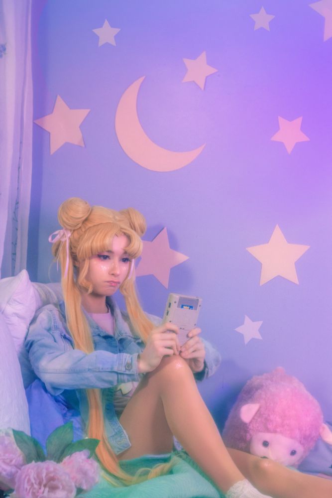 a self-taken picture of me cosplaying usagi from the anime sailor moon. i'm sitting on a bed in a denim jacket frustrated at an old gameboy from the 90s. I used saran wrap on the lens to give the photo a soft nostalgic look.