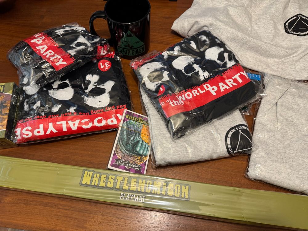 The swag on a wooden table. The swag: two sets of two different shirts, Wrestlenomicon map, cards, and a massive Delta Green Coffee mug, 