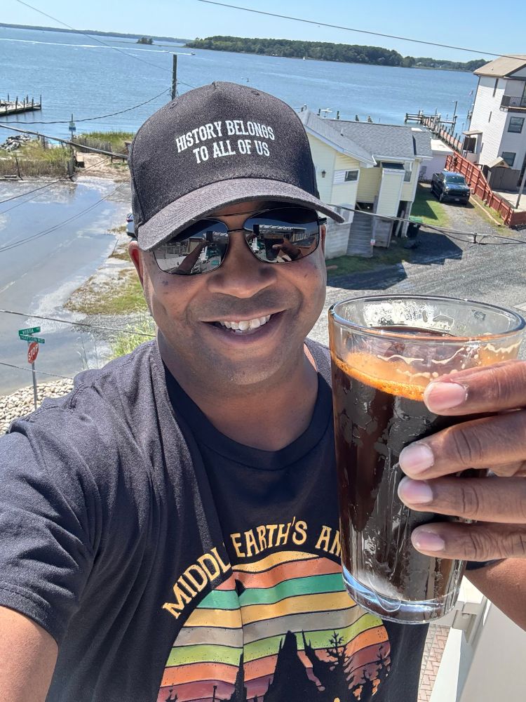 Chris holding a stout in his left hand smiling at the camera with the water behind him from the third floor. 