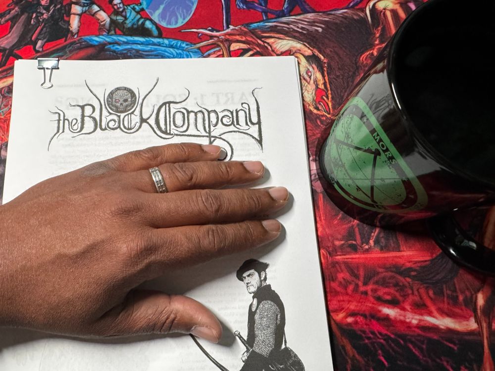 The playtest draft of Arc Dream’s The Black Company sprawls across a Haunted West mousepad, next to a half-full Delta Green coffee mug. Chris’s hand, wedding ring gleaming, partially obscures the cover and text, hinting at the work-in-progress magic within.