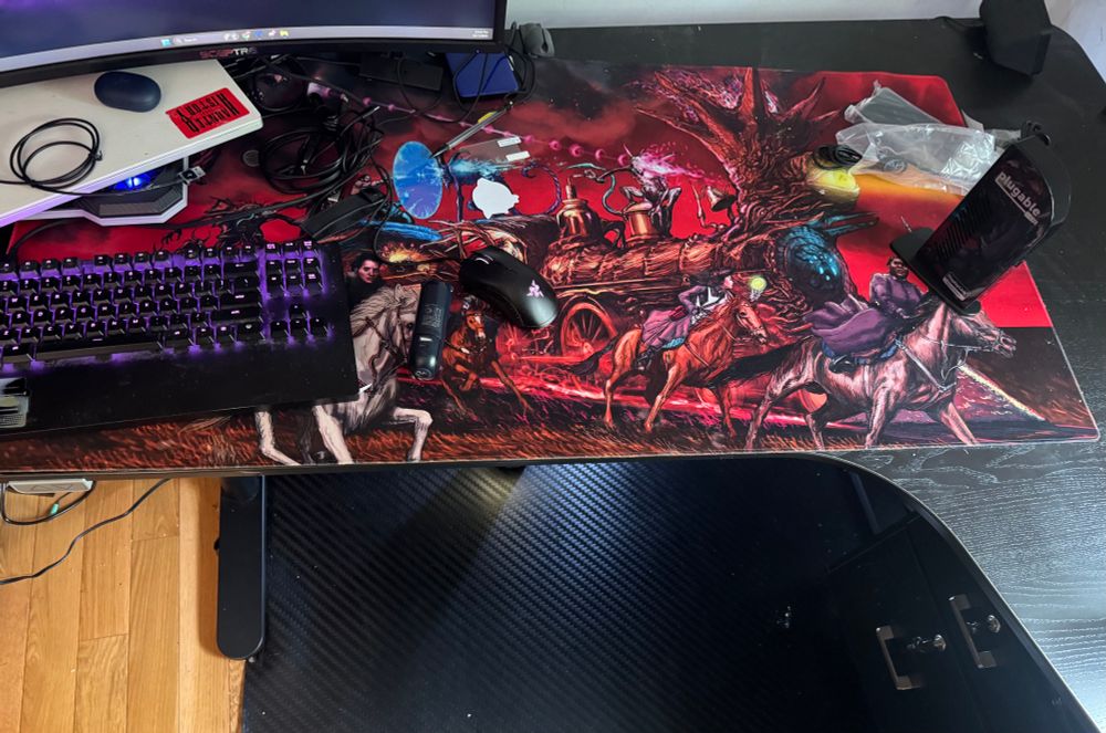 Part of Chris’s workspace stripped down with only a glowing keyboard. Long Haunted West mousepad, light illuminated under his desk and part of a laptop is visible. 