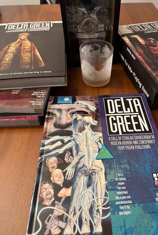 Delta Green ttrpgs new and old next to a bottle of bourbon with a frost glass in front of them with a 1 oz pour.