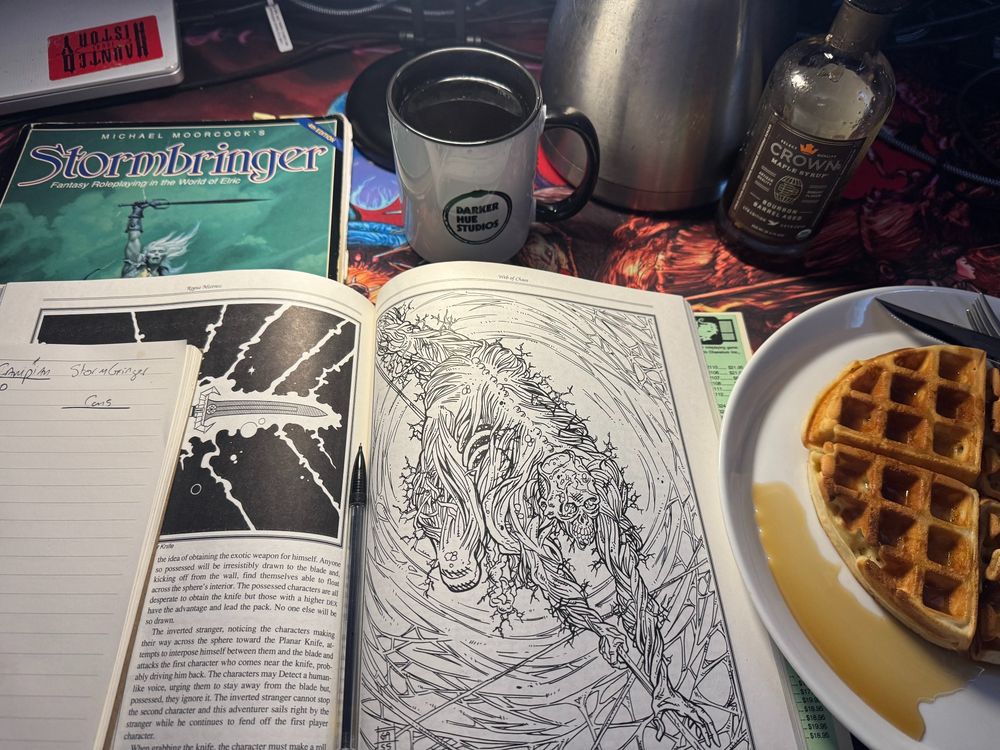 A Haunted West mousepad with a Darker Hue Studios filled coffee cup behind a Stormbringer TTRPG corebook and scenario book. Beside the books is a plate with a waffle. 